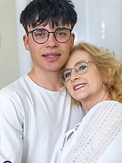 Mature cougar in leather skirt and patterned stockings embracing young man on couch