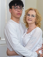 Mature cougar in leather skirt and patterned stockings embracing young man on couch